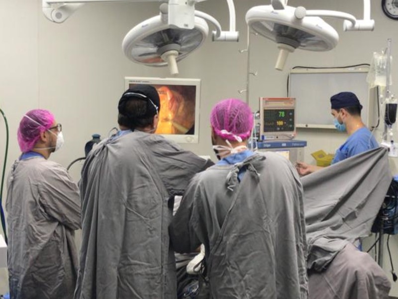 Três médicos usando roupões cinza atuam no centro cirúrgico onde é realizada uma cirurgia bariátrica. Ao centro está a mesa de operações onde o paciente está sendo operado. Há ainda um monitor de imagens e outro de batimentos cardíacos